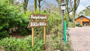 Un panneau indicateur en bois portant l’inscription « Bungalow Still Wille », entouré de verdure et surmonté d’un chemin pavé de briques menant à un bâtiment.