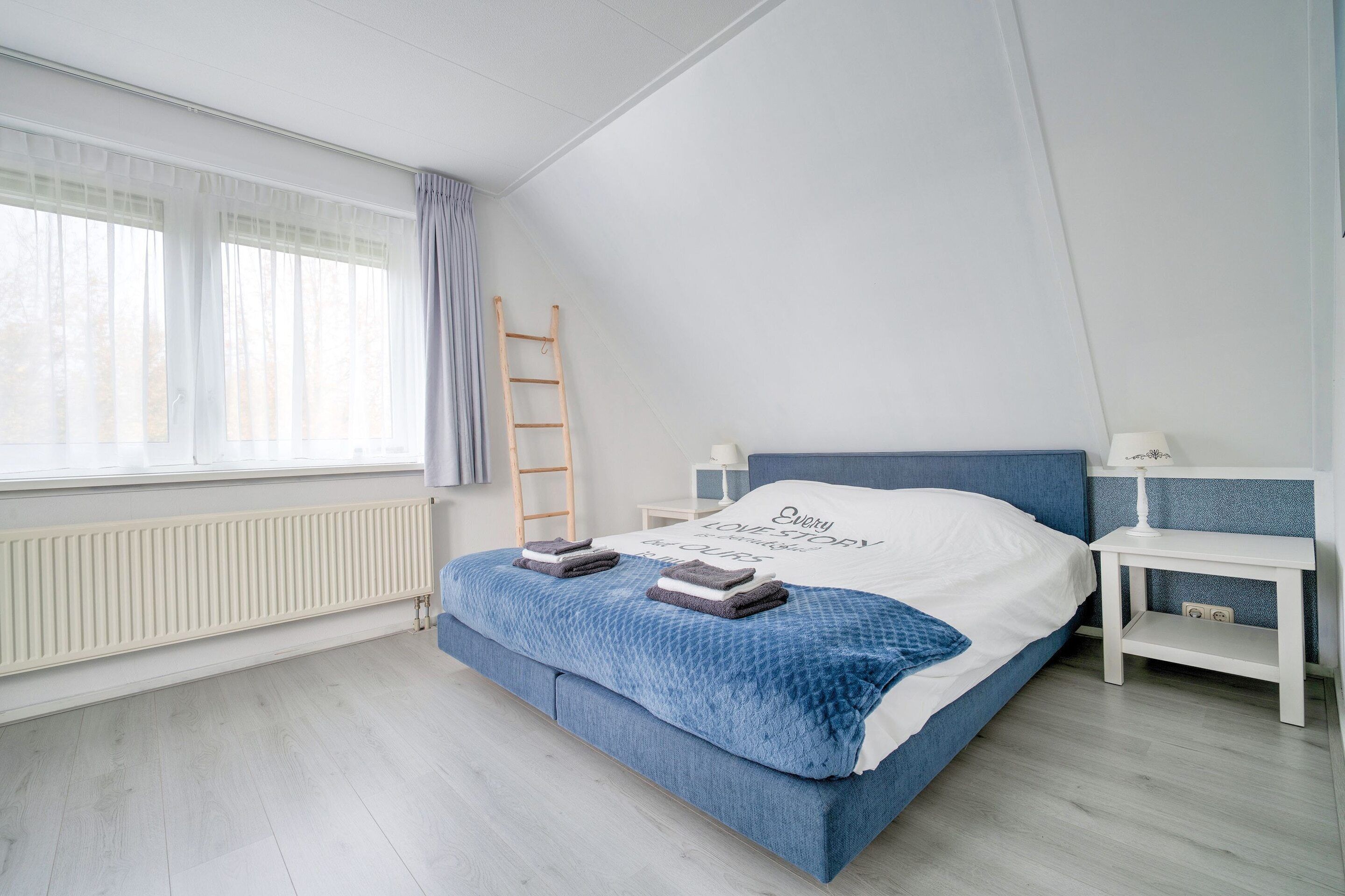 Ein Schlafzimmer mit Bett, Nachttisch, Lampe, einer Leiter und einem Fenster mit Vorhängen.