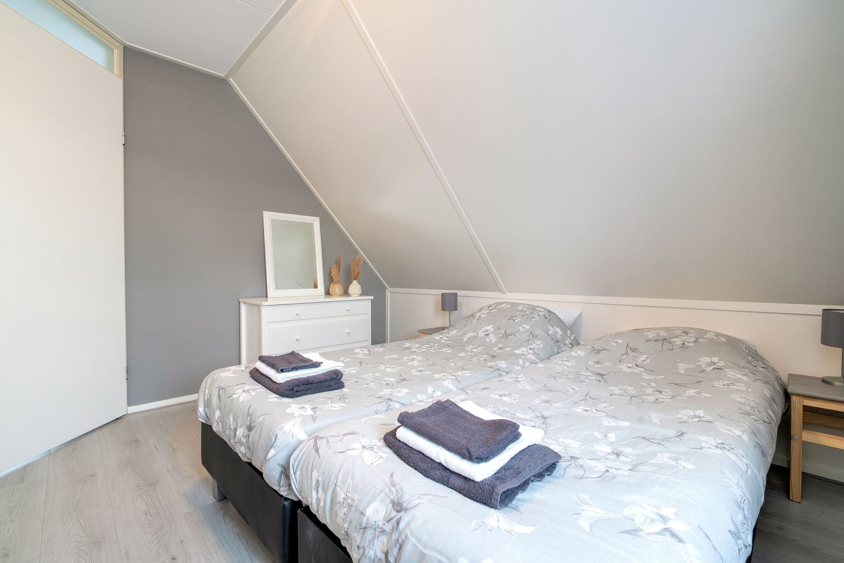 Ein Schlafzimmer mit schrägen Wänden, einem Bett mit Blumen-Überwurf, einem Nachttisch und einem Beistelltisch.