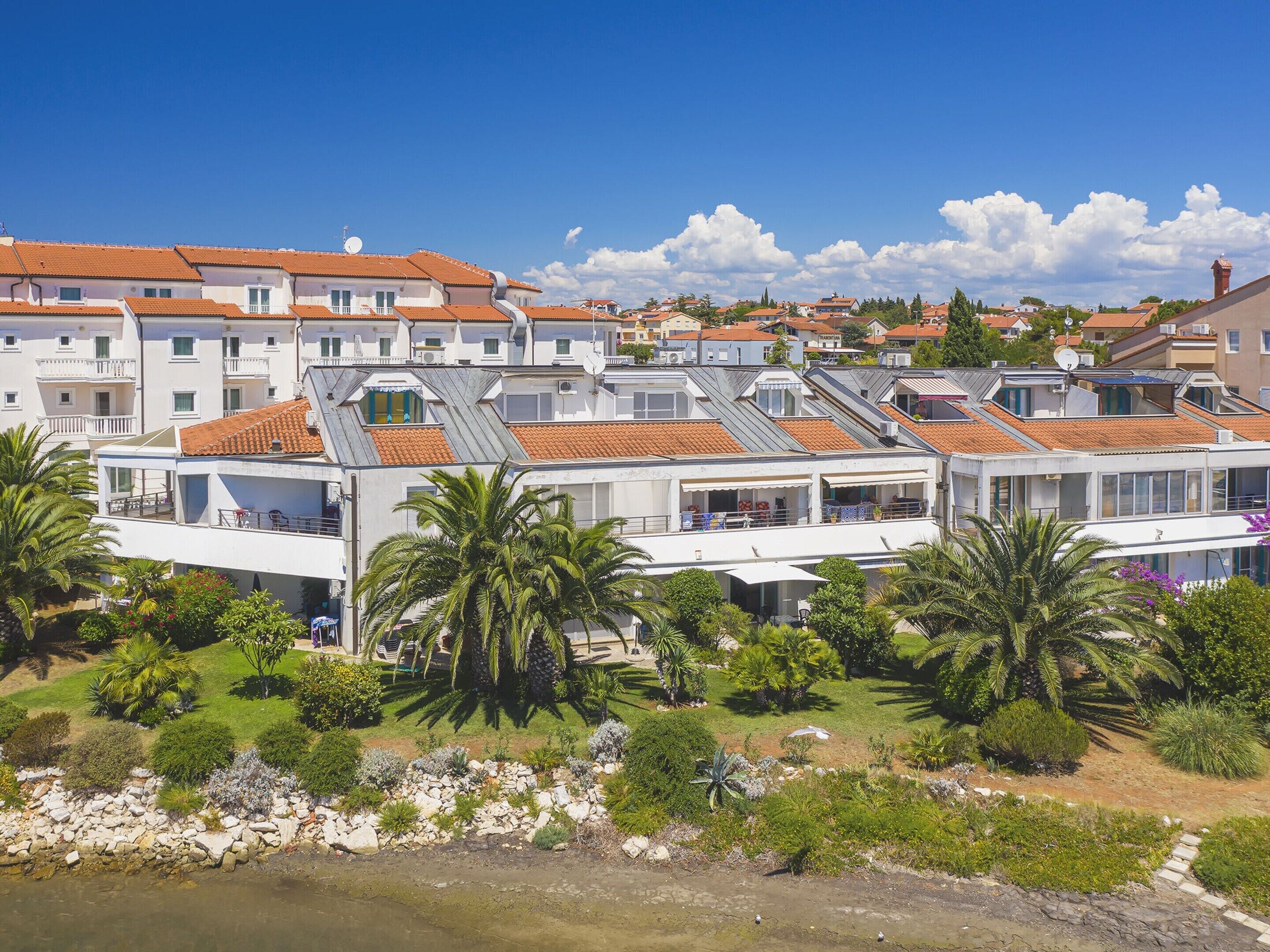 Un edificio costero con varios balcones, una terraza en la azotea y un jardín con palmeras.