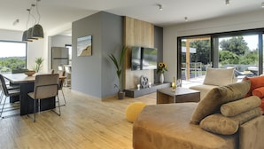 A modern living room with a large sofa, a dining area with a table and chairs, a TV mounted on the wall, and a view of greenery outside. - Comfortable, Stylish, Detached Villa With Garden, Private Pool, Near the Beach (Medulin)