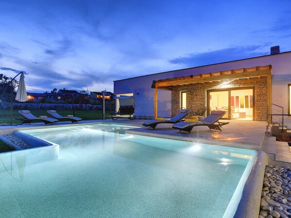 A modern outdoor pool area with lounge chairs and a stone building with a glass door.