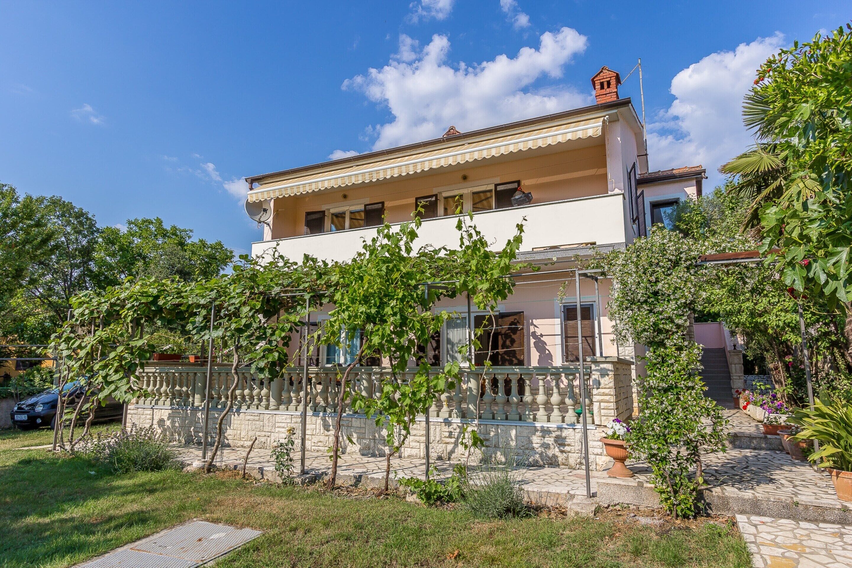 A two-story building with a balcony, surrounded by greenery and a stone pathway.