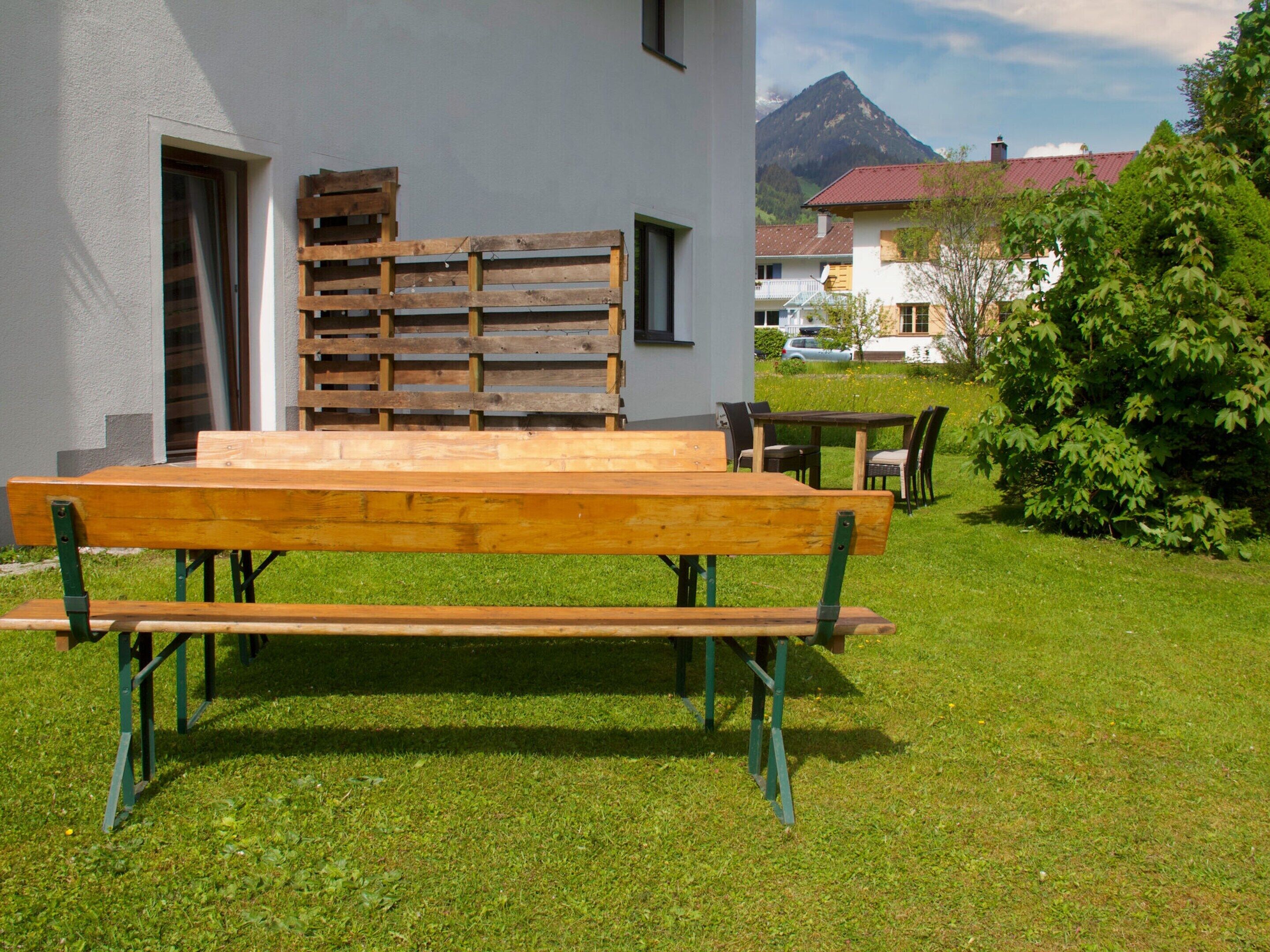 Un conjunto de banco y mesa de madera al aire libre frente a un edificio, con una montaña de fondo.