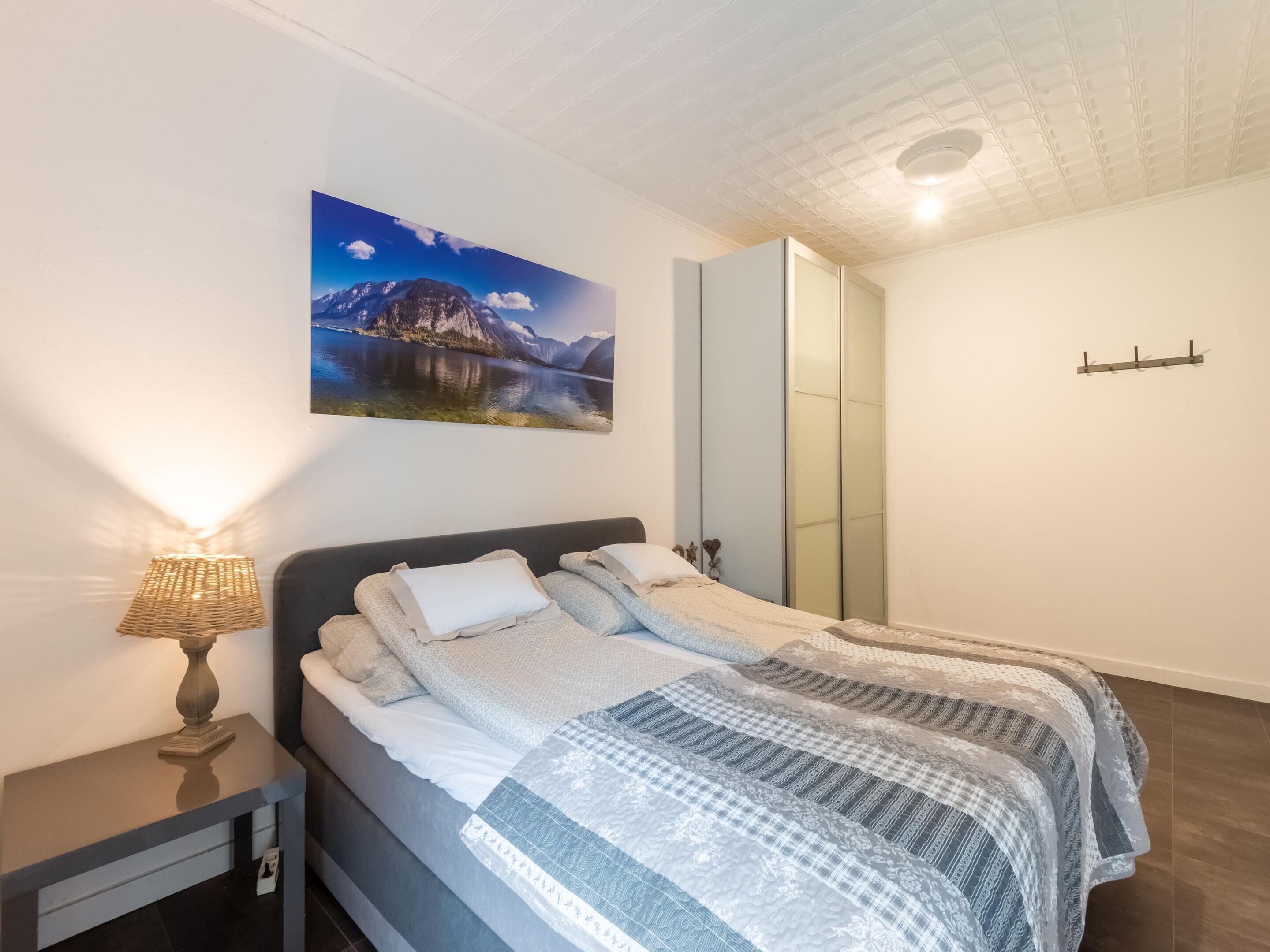 Une chambre à coucher comprenant un lit, une table de chevet, une lampe, un tableau représentant un paysage de montagne et une armoire.
