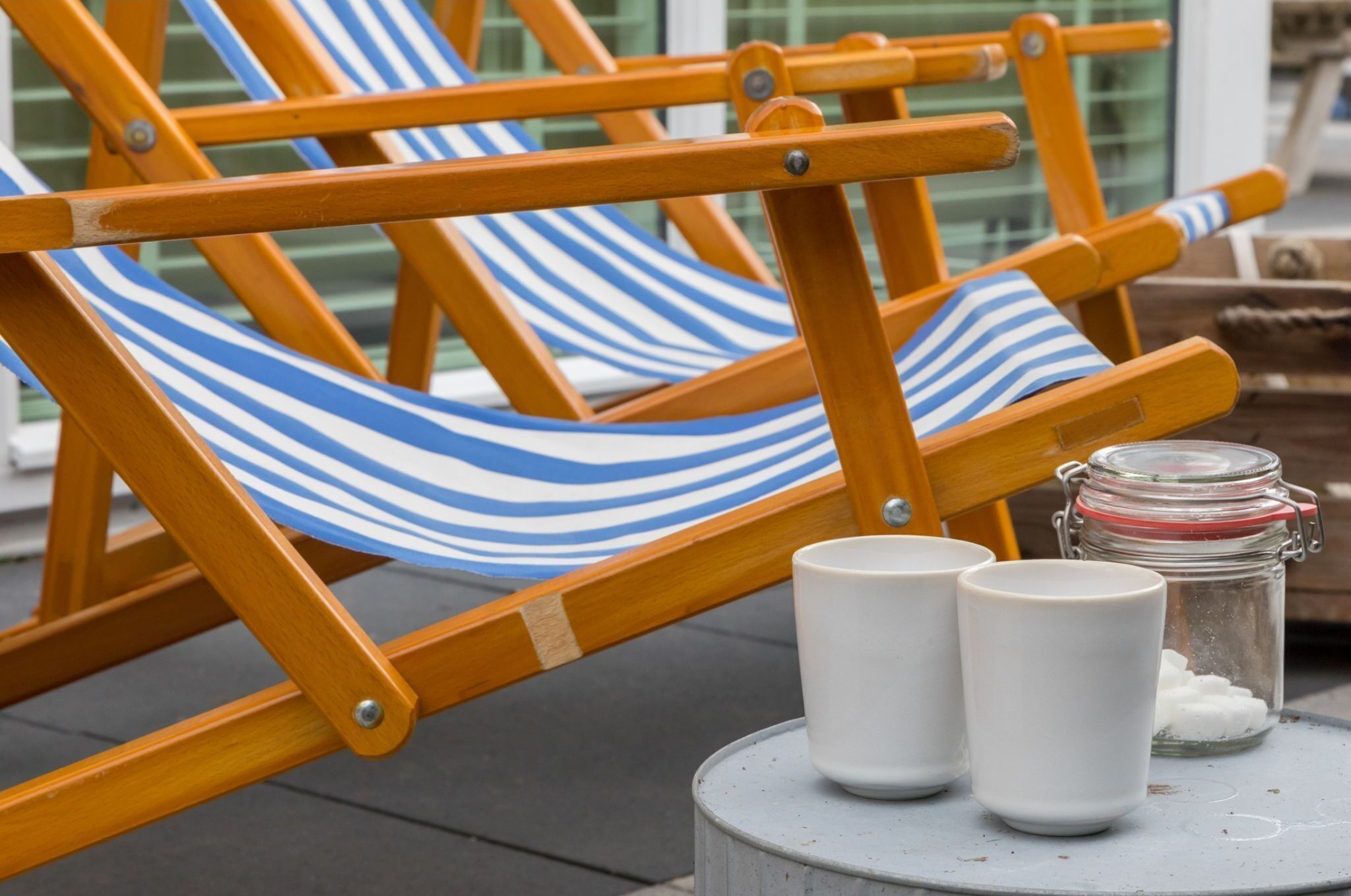 A wooden chair with a blue and white striped hammock, two white ceramic cups, and a jar with sugar on a round table.