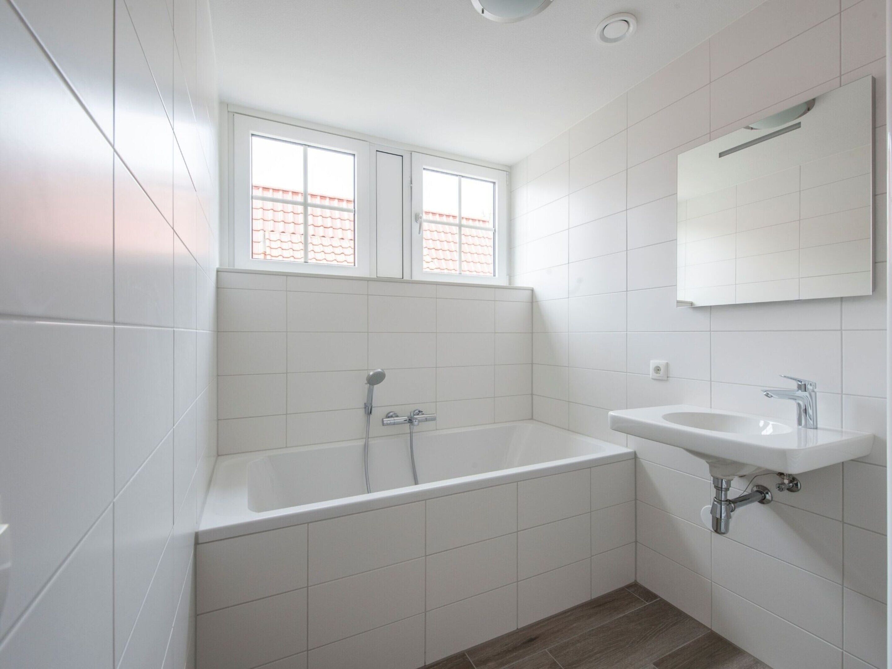 Een moderne badkamer met witte tegels, een badkuip, een wastafel en een raam met uitzicht op een dak met rode tegels.
