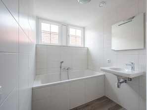 Un bagno moderno con piastrelle bianche, una vasca da bagno, un lavandino e una finestra con vista su un tetto in tegole rosse.