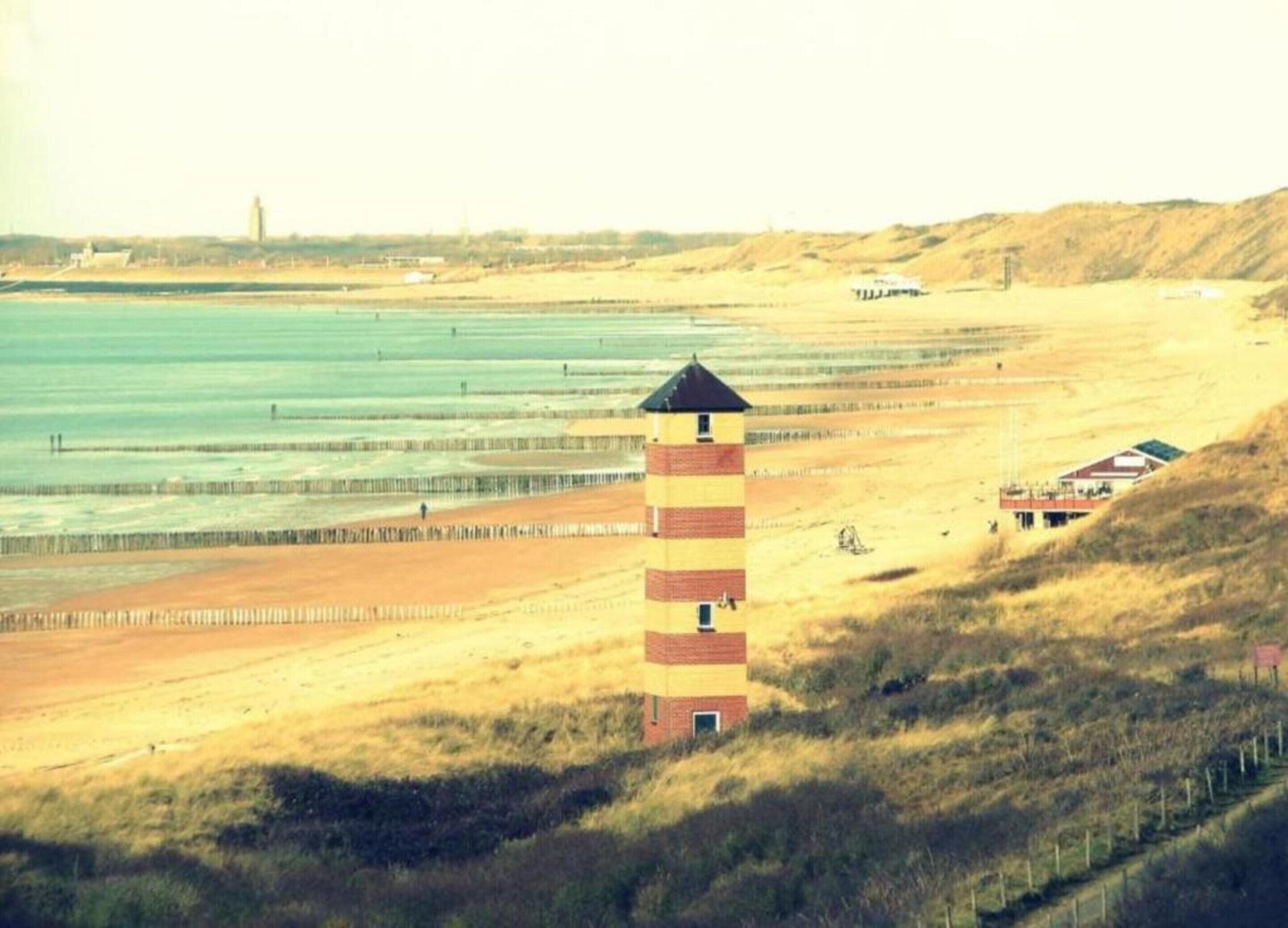 Eine Küstenlandschaft mit einem markanten Leuchtturm, einem Sandstrand und einem kleinen Gebäude mit rotem Dach.