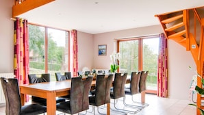 A dining area with a wooden table and chairs, large windows with curtains, and a staircase.