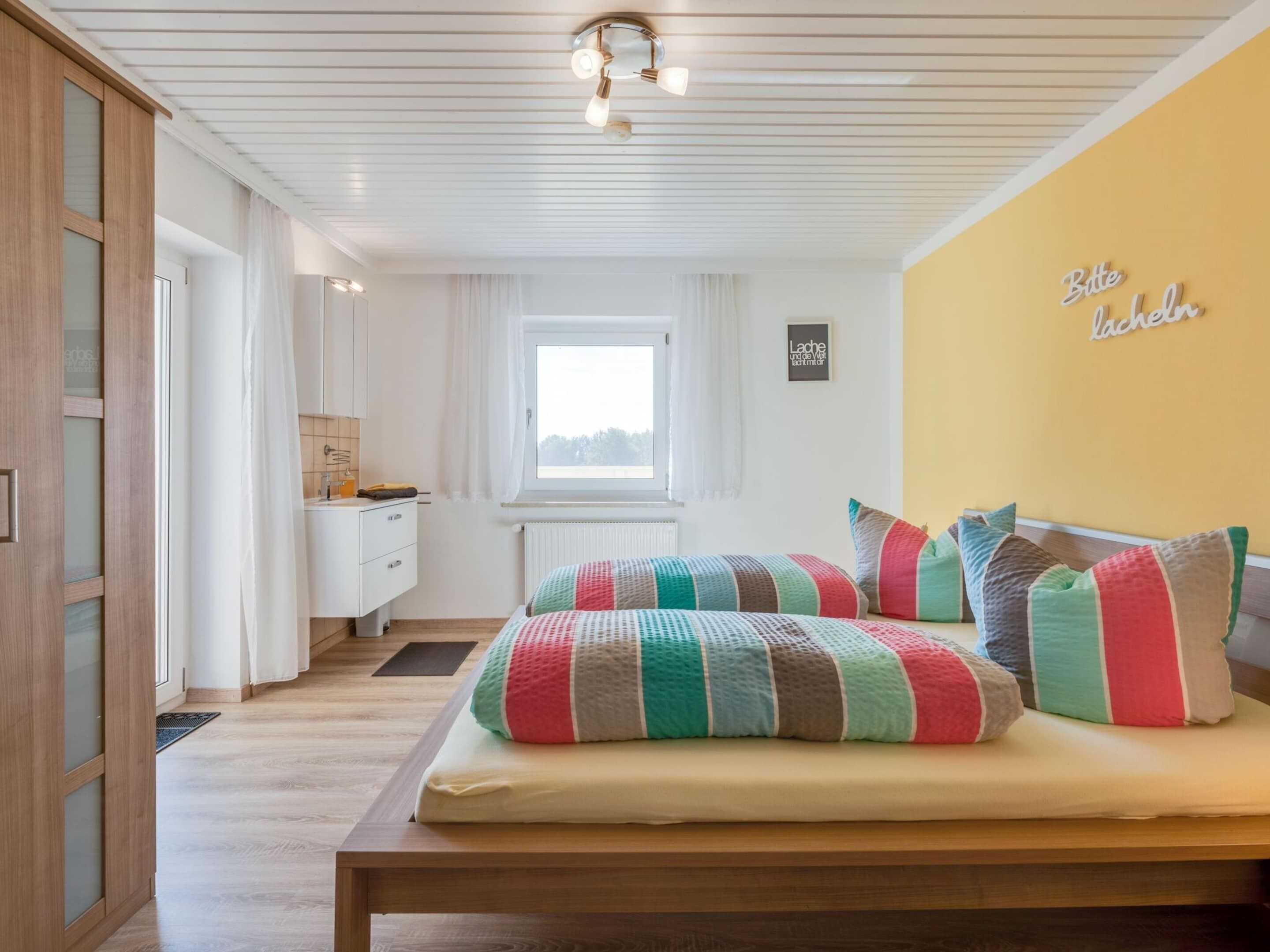 Een slaapkamer met een houten bed, kleurrijke kussens, een raam, een witte garderobe en een muur met een bord.