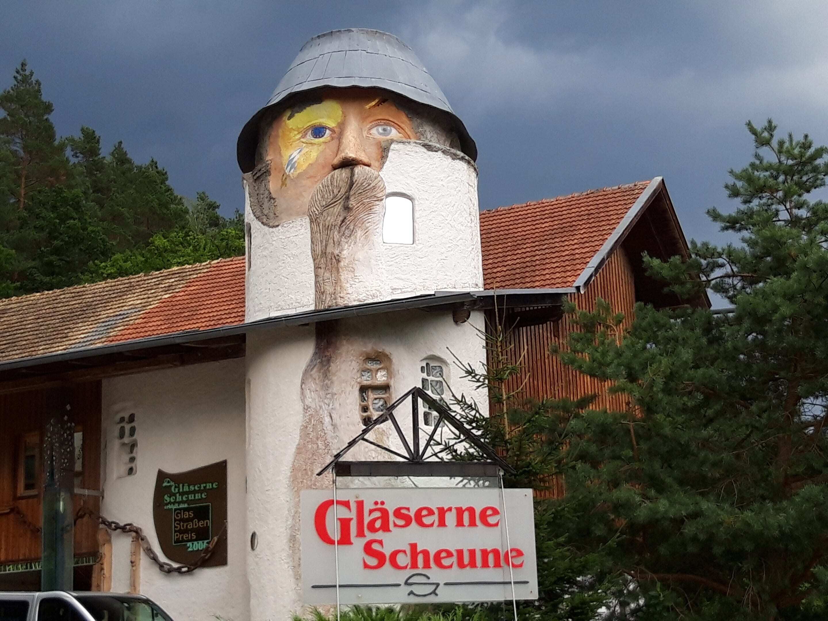 A building with a large mural on its side, a sign reading "Gläserne Scheune," and a wooden structure with a red roof.
