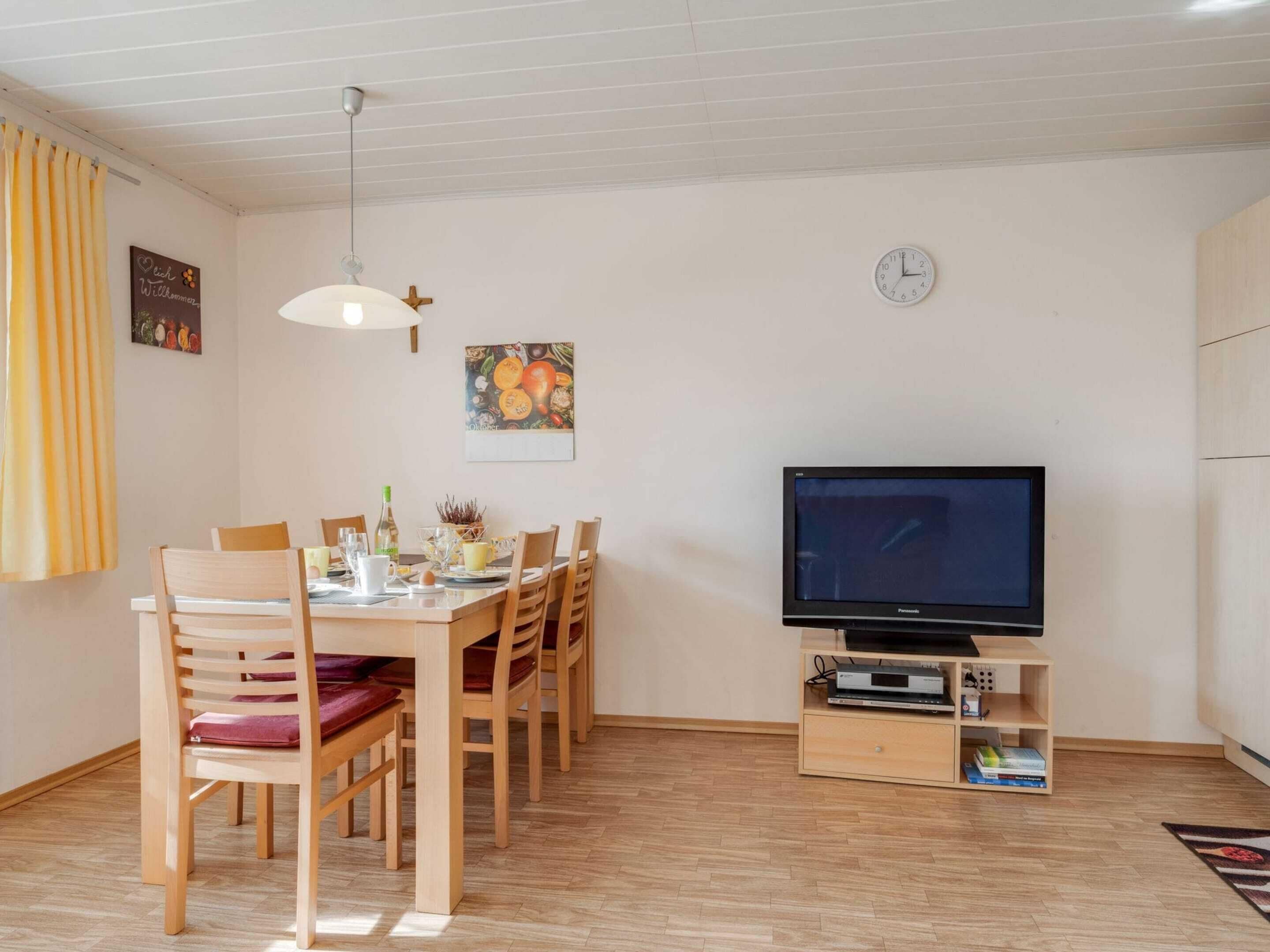 A dining area with a wooden table and chairs, a television on a stand, and a clock on the wall.