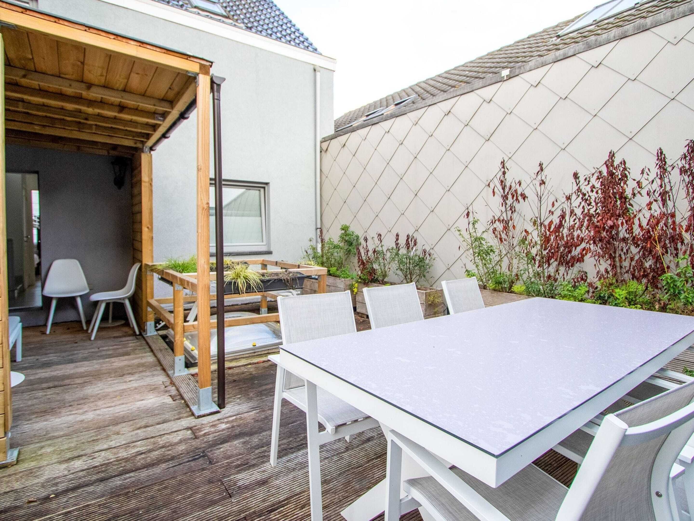 Eine Terrasss mit Tisch und Stühlen, einer Holz-Pergola und einem Garten mit Pflanzen. Eine Wand zeigt ein Rautenmuster.