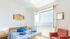 A bedroom with a wooden bed, a red chair, a nightstand with a lamp, and a window with a view of the sea. - Sprawling Apartment in Cala Gonone near Cala Fuili Beach (Dorgali)