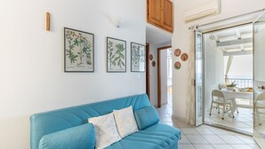 A living room with a blue sofa, framed botanical prints, a wooden cabinet, and a glass door leading to a balcony with outdoor furniture. - Sprawling Apartment in Cala Gonone near Cala Fuili Beach (Dorgali)