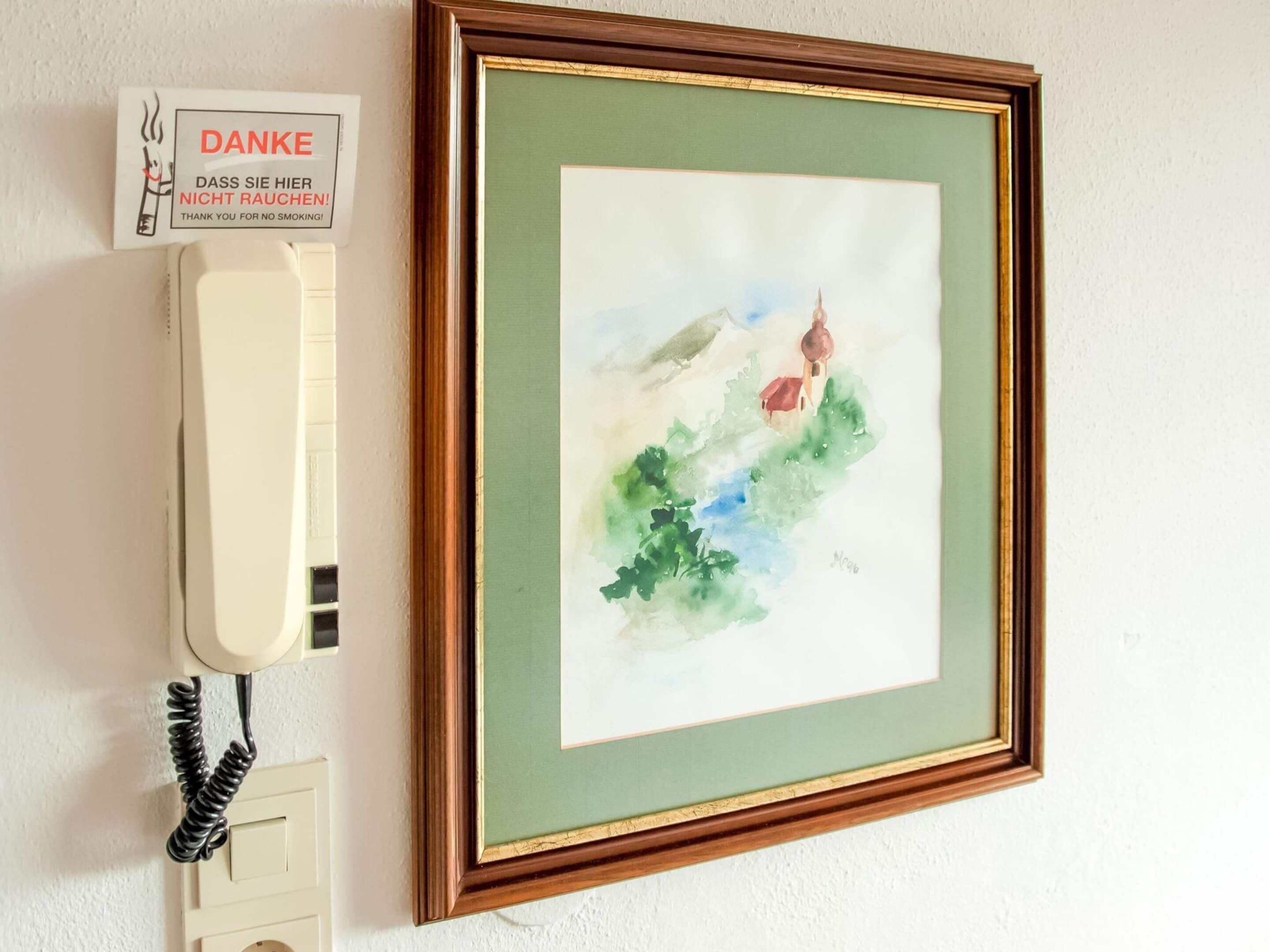 Pintura em aquarela emoldurada retratando uma paisagem com uma casa e árvores, fixada numa parede ao lado de um telefone.