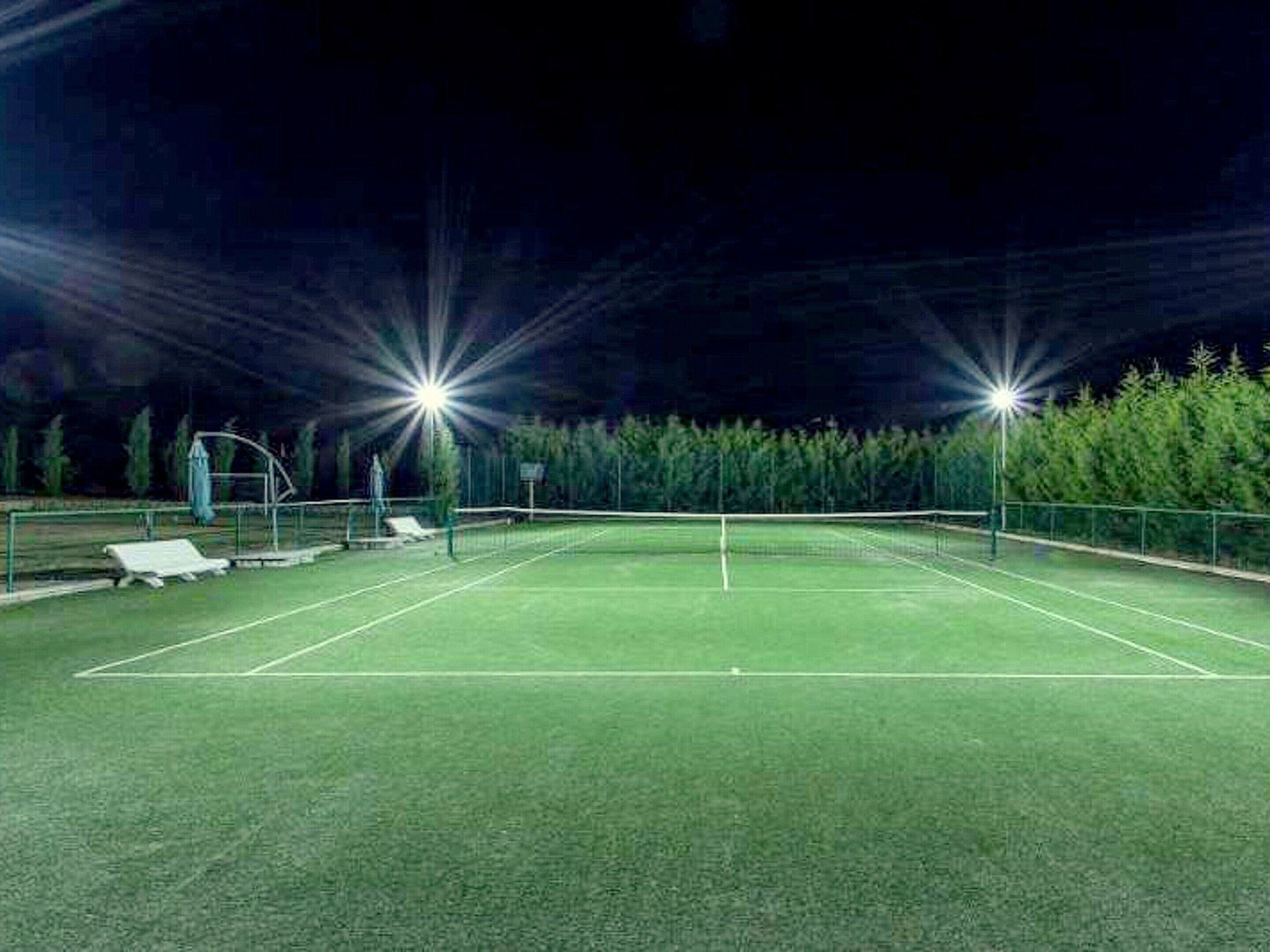 Ein Tennisplatz im Freien bei Nacht mit Flutlicht, umgeben von Bäumen.