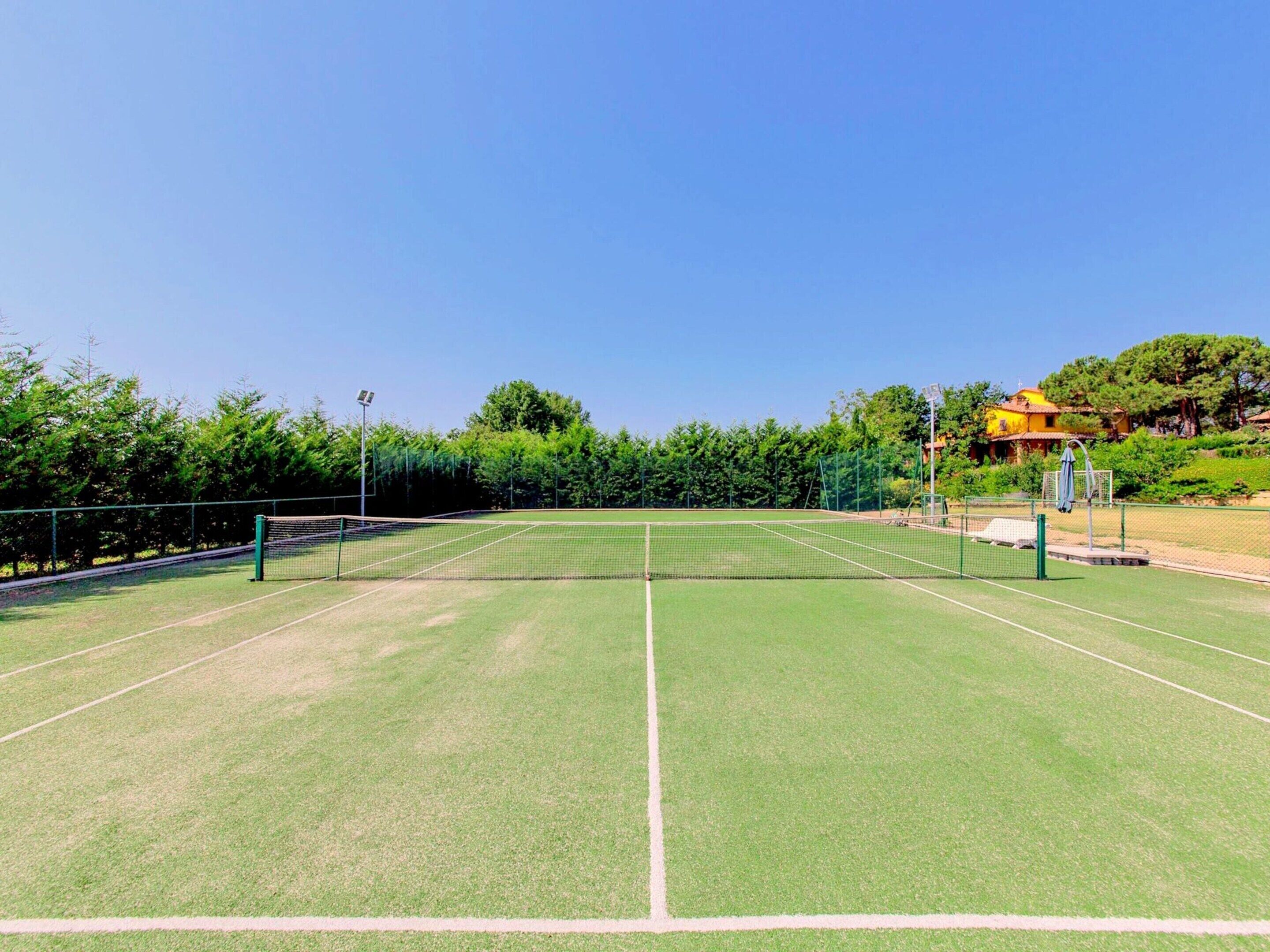Un court de tennis avec un filet, entouré de verdure et avec un bâtiment jaune en arrière-plan.