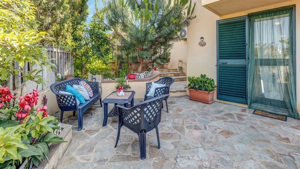 Un patio al aire libre con suelo de piedra, dos sillas de mimbre negro, una mesita y plantas en macetas.