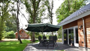 An outdoor patio with a table and chairs under a large umbrella, surrounded by trees and a wooden building with a glass door. - House in Reutum With Jacuzzi & Forest View (Reutum)