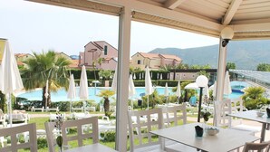 Area pranzo all'aperto con tavoli e sedie bianche, con vista su una piscina e un paesaggio montuoso.