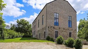 A two-story stone building with a pitched roof, surrounded by greenery and a gravel driveway.