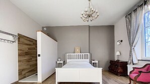 A bedroom with a bed, a wooden wardrobe, a dresser, a chair, a chandelier, and a window with curtains.