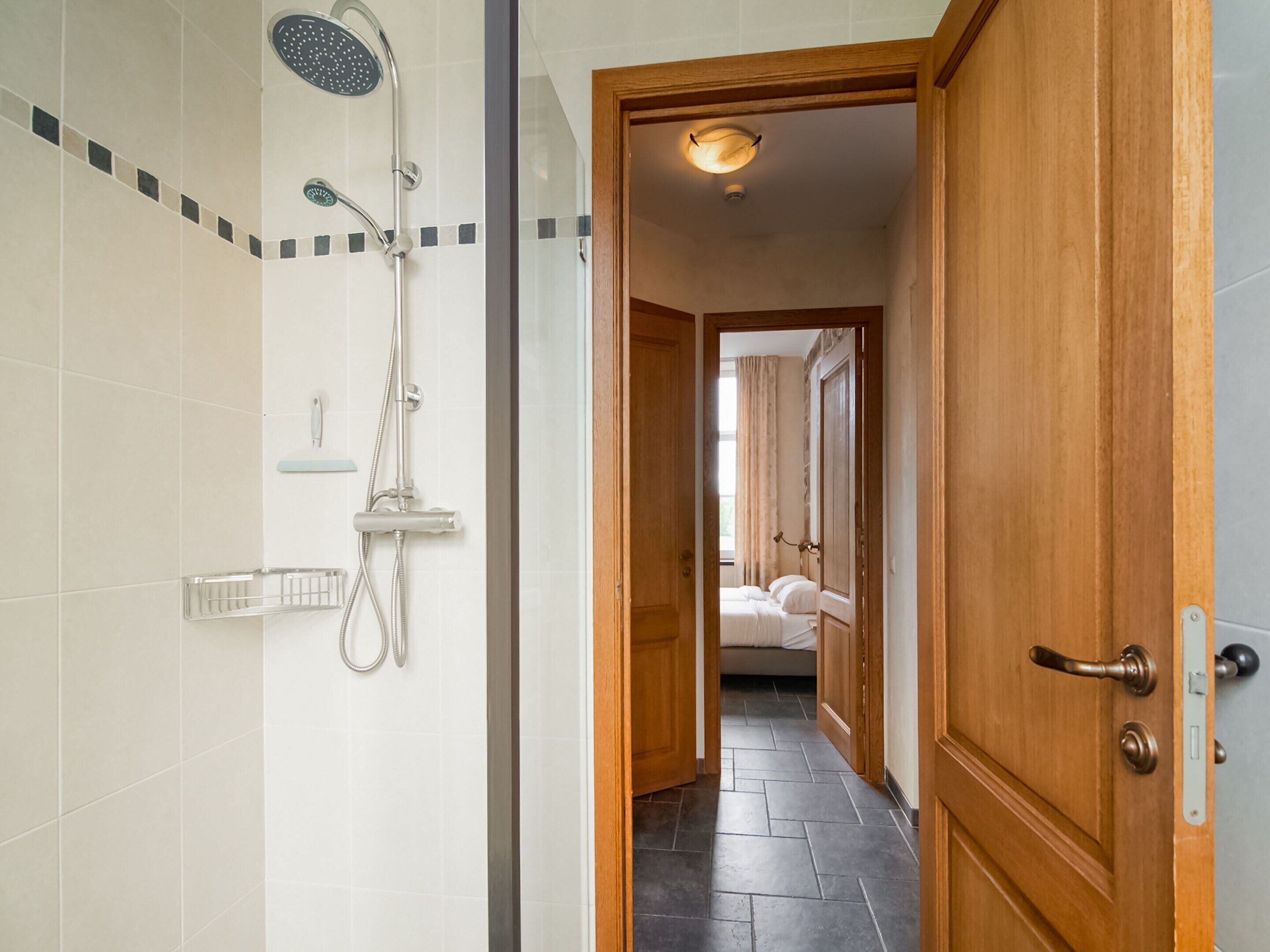 A hotel bathroom with a shower, toilet, and sink. A wooden door leads to a bedroom with a bed and window.