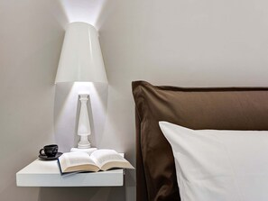 Un lit bien fait, avec une tête de lit marron, une table de chevet blanche sur laquelle se trouve une lampe, un livre ouvert et une tasse noire.