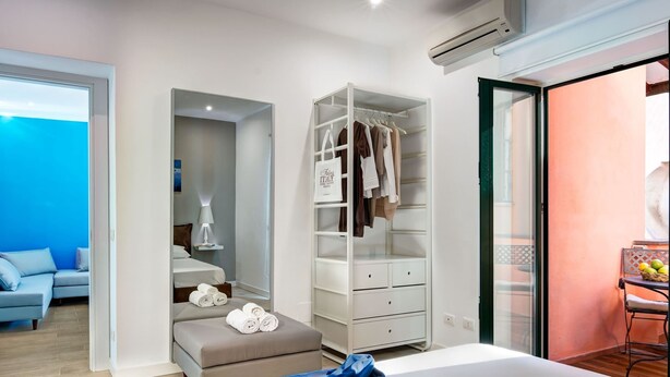 Una habitación de hotel moderna con una cama, un vestidor, una zona de estar con una mesa y sillas, y vistas a través de una puerta abierta.