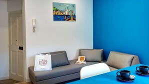 Una habitación con pared azul, un sofá con cojines, una mesa con dos tazas y platos, un cuadro de una escena costera y un bolso blanco sobre el sofá.