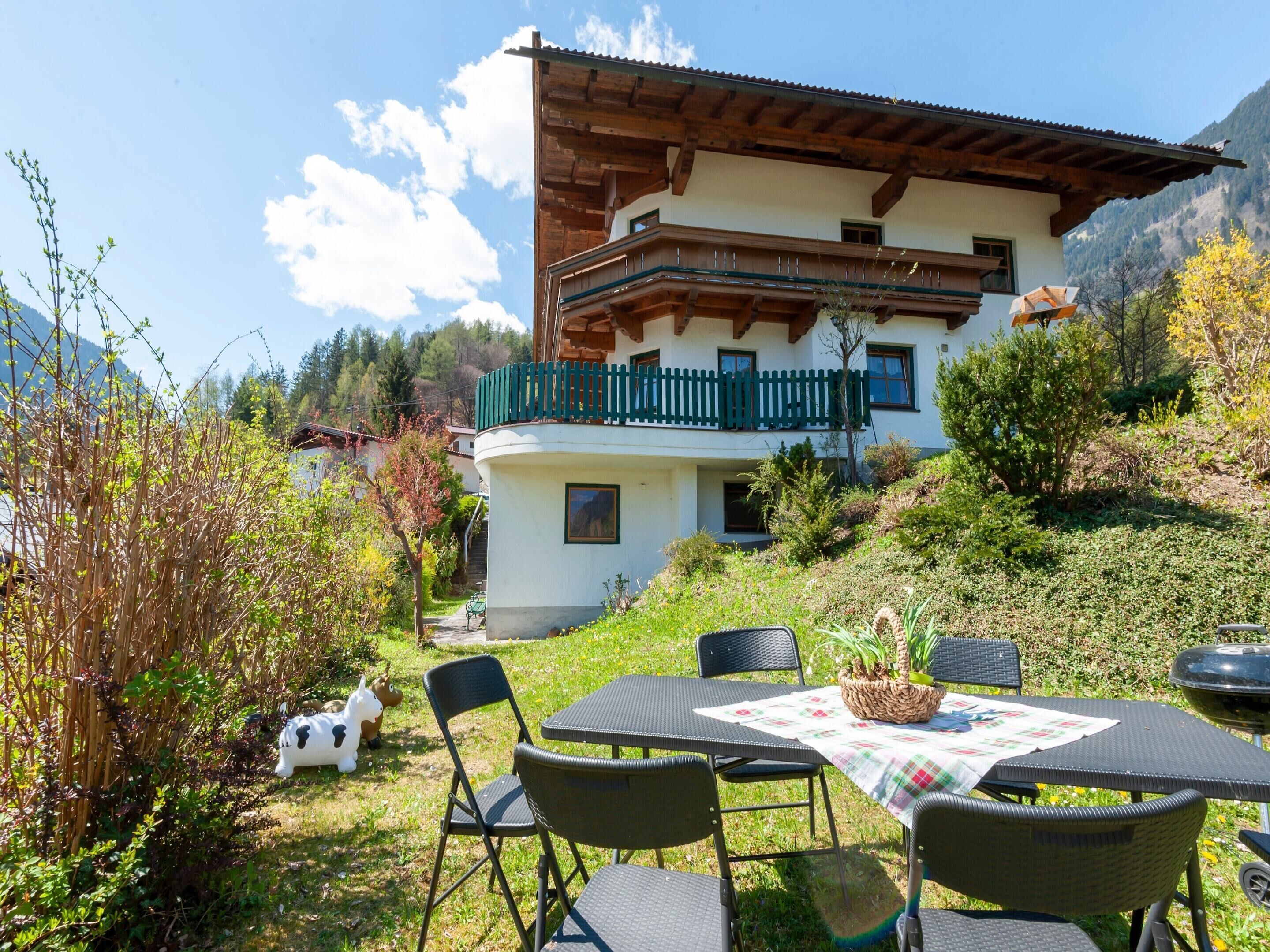 A two-story chalet with a balcony, surrounded by lush greenery and mountains in the background.
