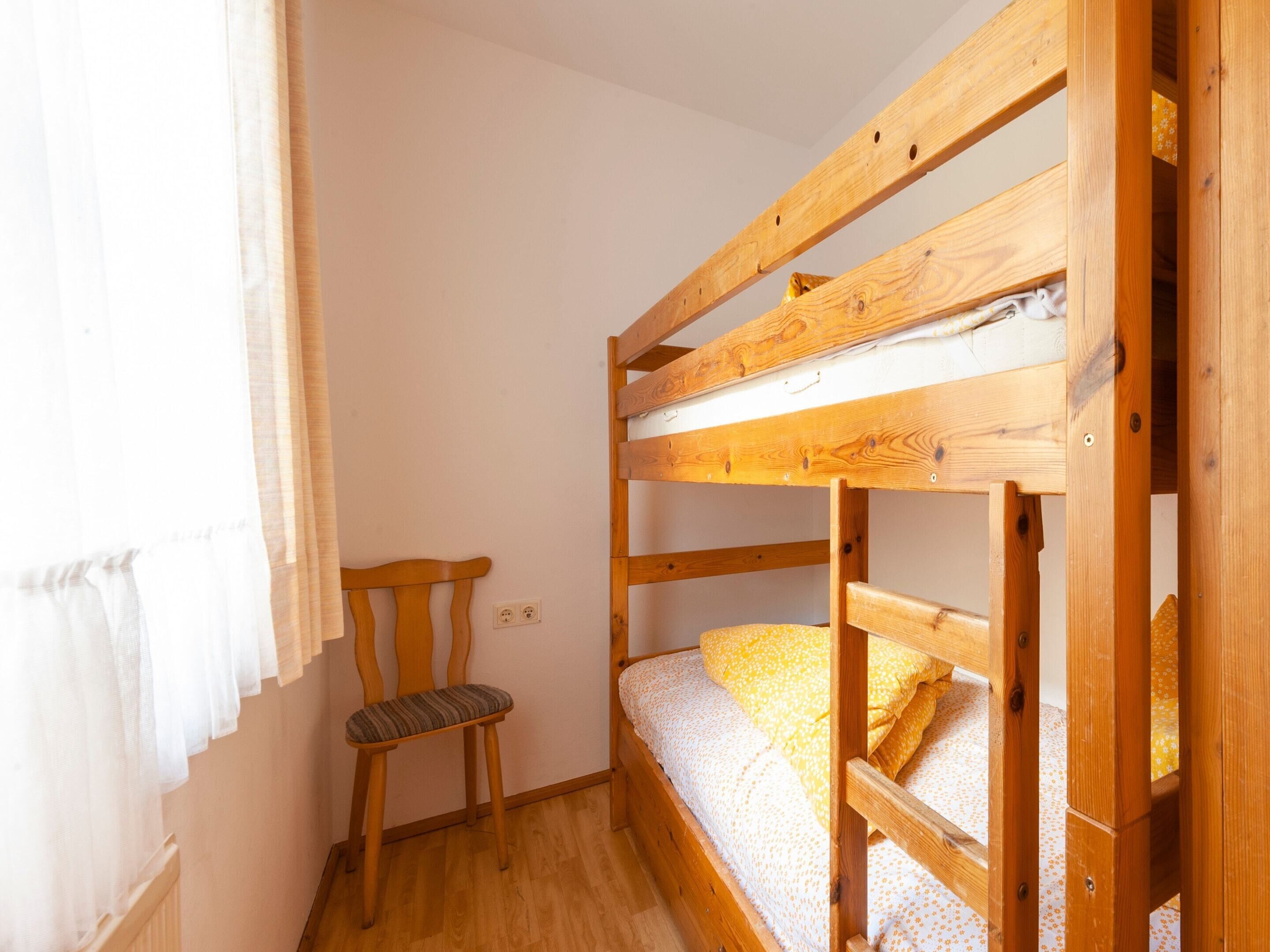 A bunk bed with a wooden frame, a chair with a striped cushion, and a window with white curtains.