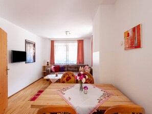Una sala de estar con un sofá, un televisor montado en la pared, una mesa de comedor con camino de mesa floral y una ventana con cortinas.