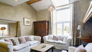 A spacious living room with a high ceiling, a wooden ceiling beam, a large window with curtains, a wooden cabinet, and a small table with decorative items. - Magnificent Holiday Home in Manhay With Fenced Garden (Manhay)
