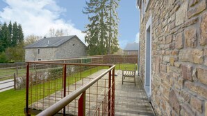 A wooden deck with a metal railing, a stone building, and a grassy area with trees. - Magnificent Holiday Home in Manhay With Fenced Garden (Manhay)