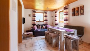 Ein gemütliches Zimmer mit Holzbalkendecke, einer Couch, einem Esstisch mit Hocker und einem Fenster mit Vorhängen.
