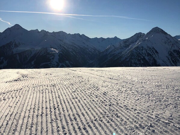 A snow-covered ski slope with ski tracks, surrounded by mountains under a clear sky.