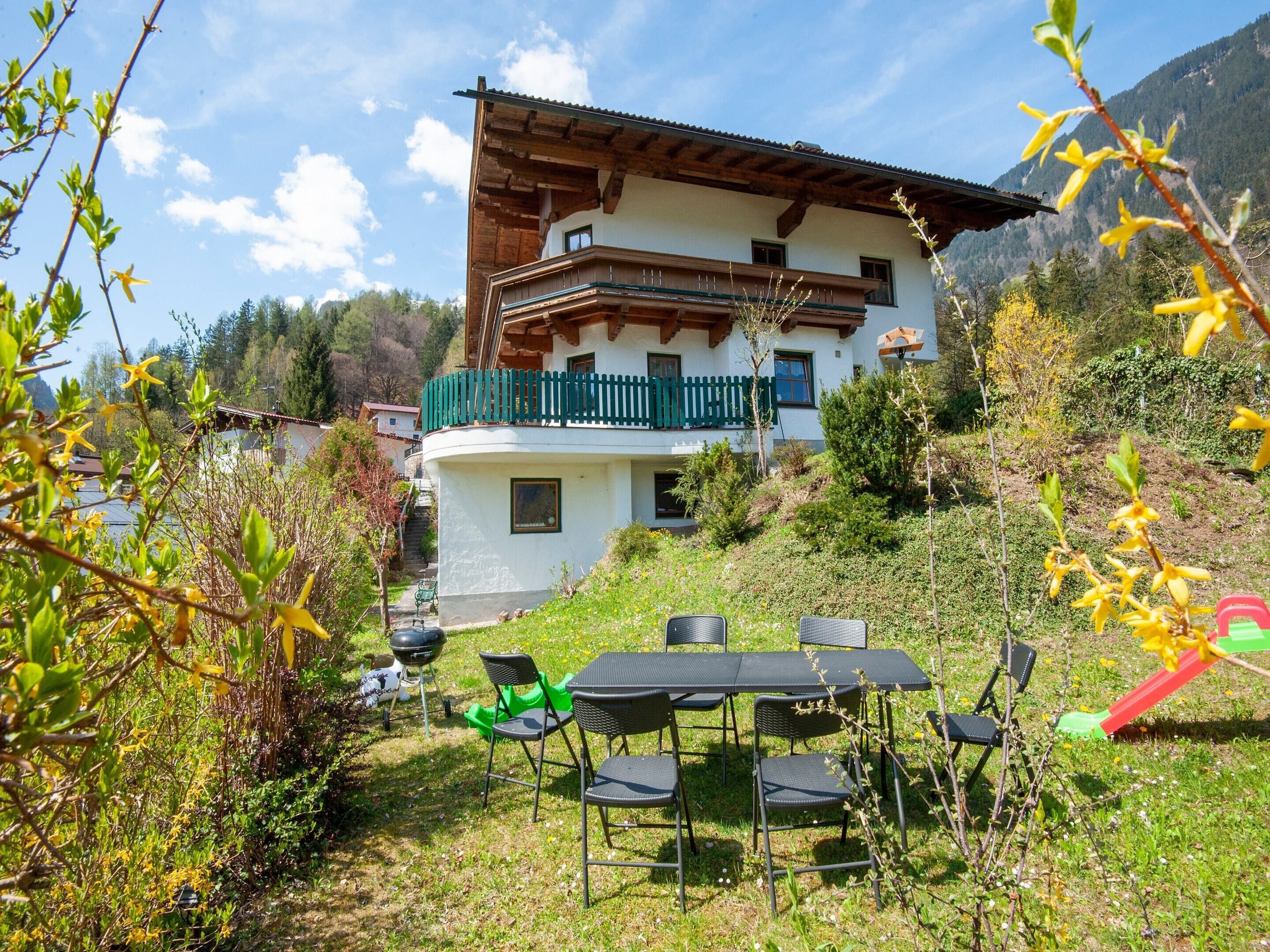 A two-story chalet with a balcony, surrounded by greenery and a mountain backdrop.