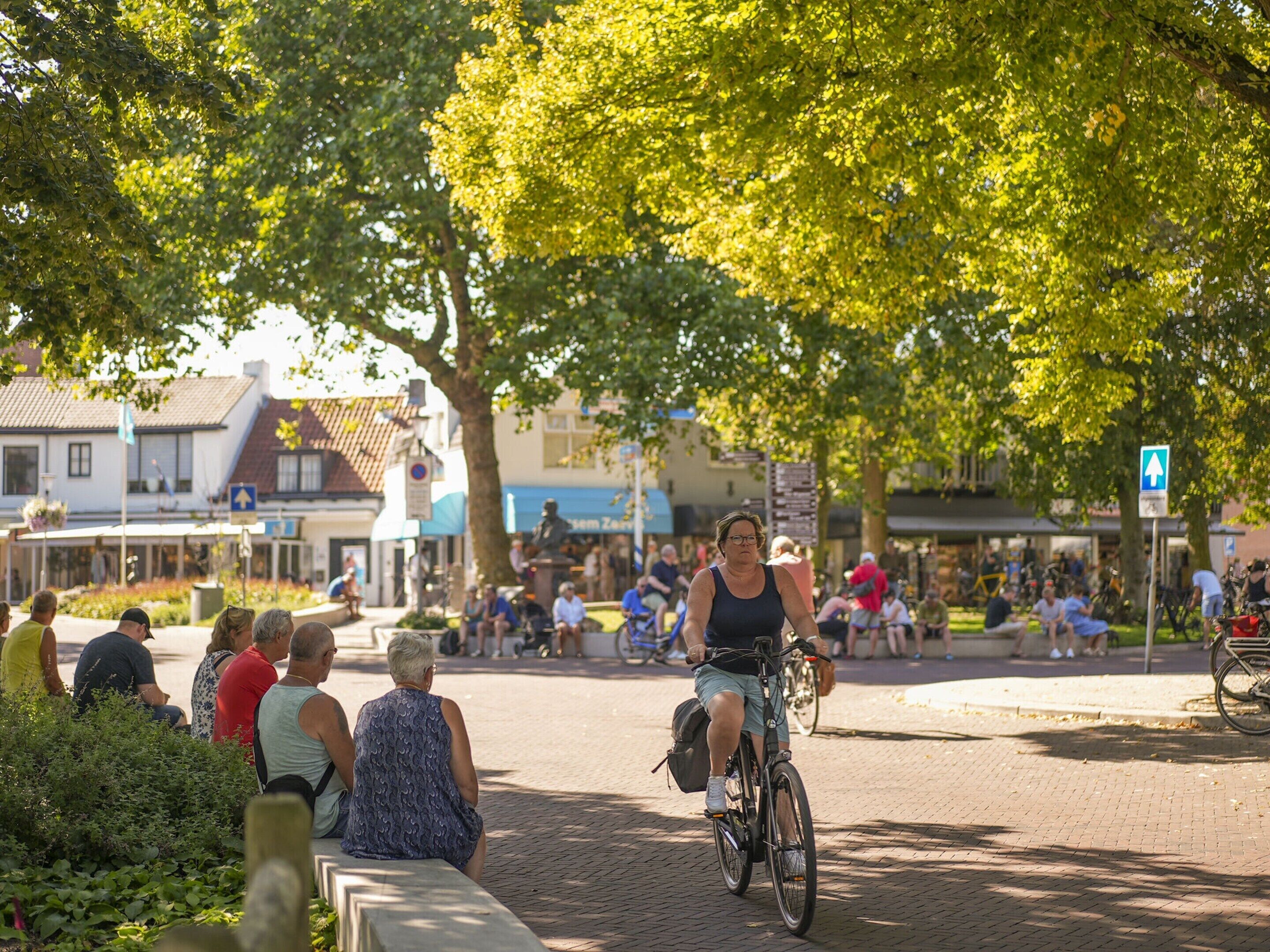 A cyclist riding on a paved path in a public square with people sitting on benches and others walking around.