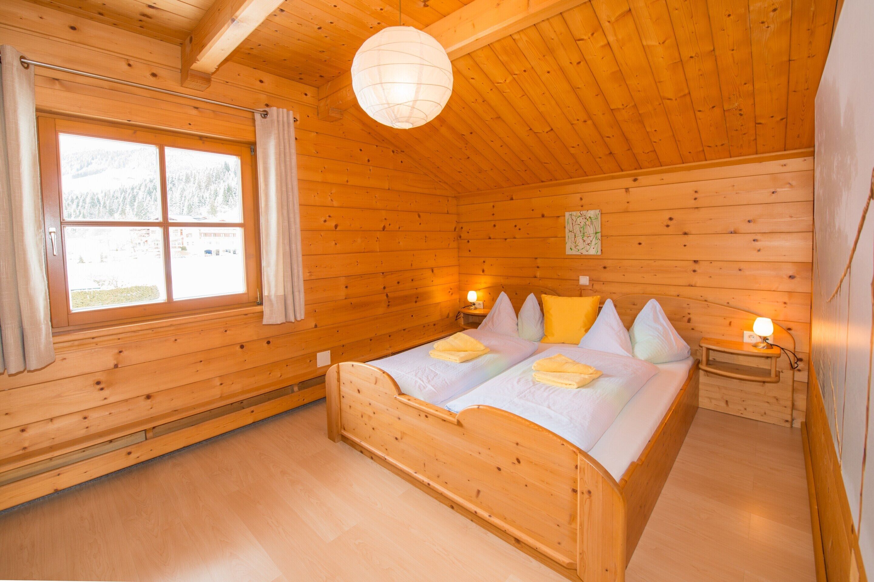 A wooden cabin bedroom with a bed, two bedside tables, a hanging light fixture, and a window with curtains.