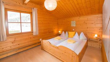 A wooden cabin bedroom with a bed, two bedside tables, a hanging light fixture, and a window with curtains.