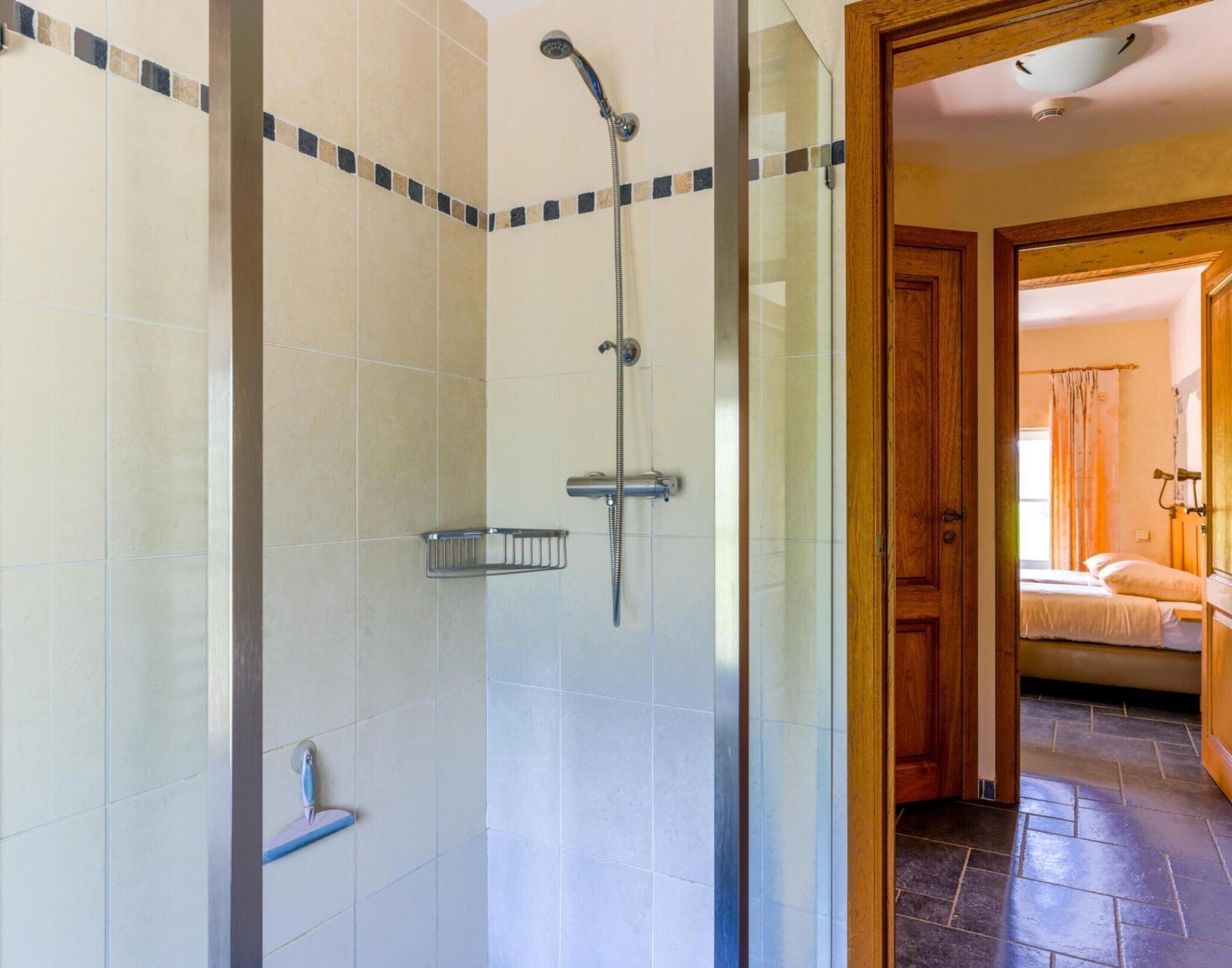 A shower area with a handheld showerhead and a wall-mounted faucet. There is a small metal shelf attached to the wall.