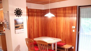 A dining area with a wooden table, two chairs, a red cushion, a wall clock, and a hanging light fixture. - Serene Apartment in Bohinjsko Jezero near Ski Lift (Bohinj)
