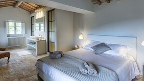 A bedroom with a large bed, a bedside table with a lamp, a window, a bathroom with a sink, and a chair.