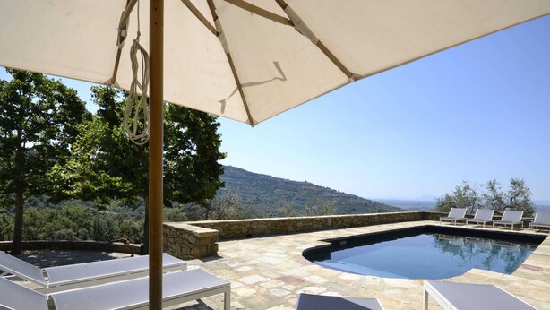 A pool area with lounge chairs and an umbrella, surrounded by stone walls and trees, overlooking a hilly landscape.