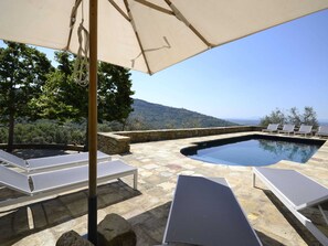 A pool area with lounge chairs and an umbrella, surrounded by stone walls and trees, overlooking a hilly landscape. - Belvilla by OYO Pomona (Cortona)