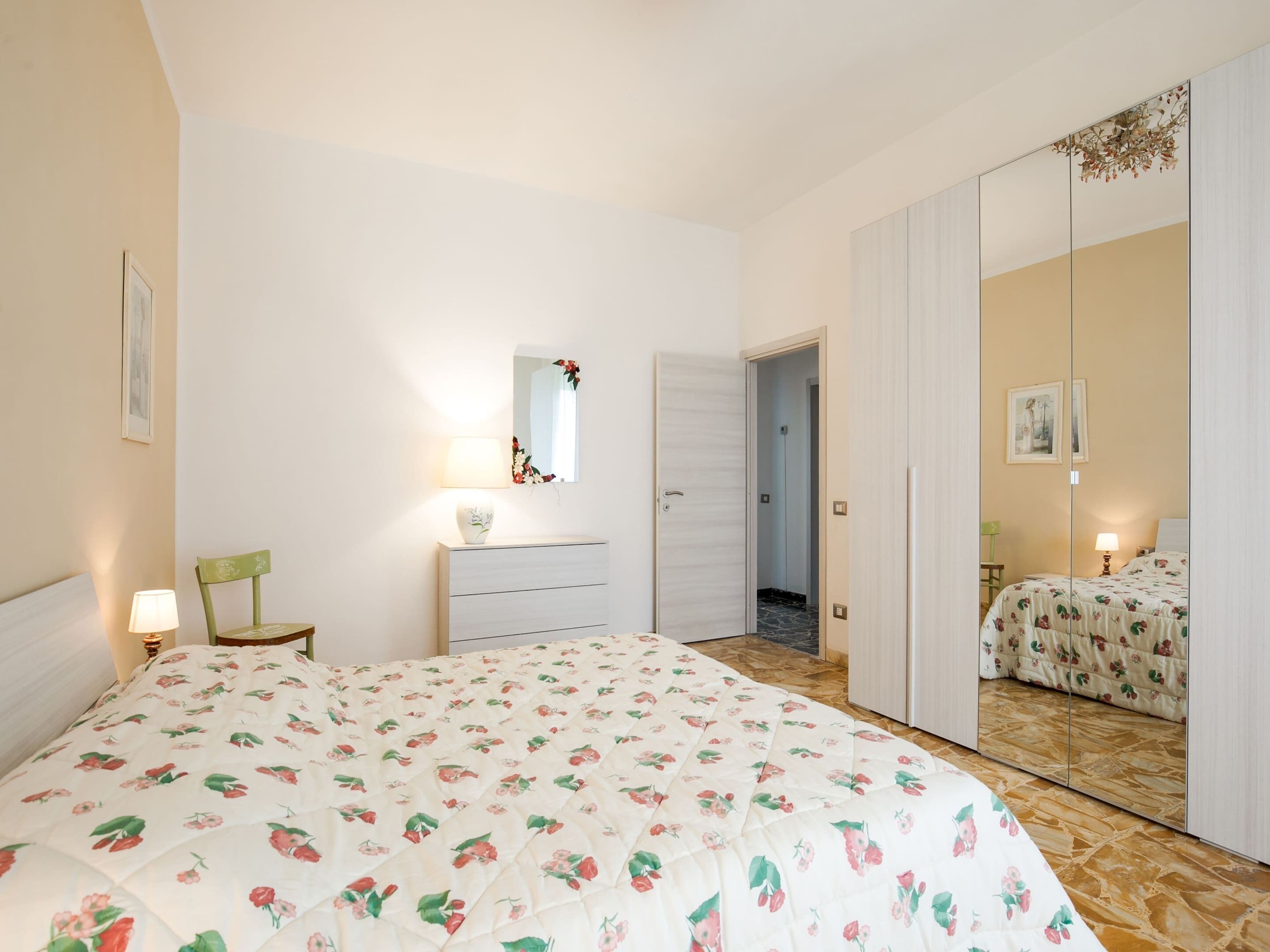 Une chambre avec une couvre-lit à motifs floraux, une commode avec un miroir, une porte donnant sur une autre pièce et une armoire à portes miroir.