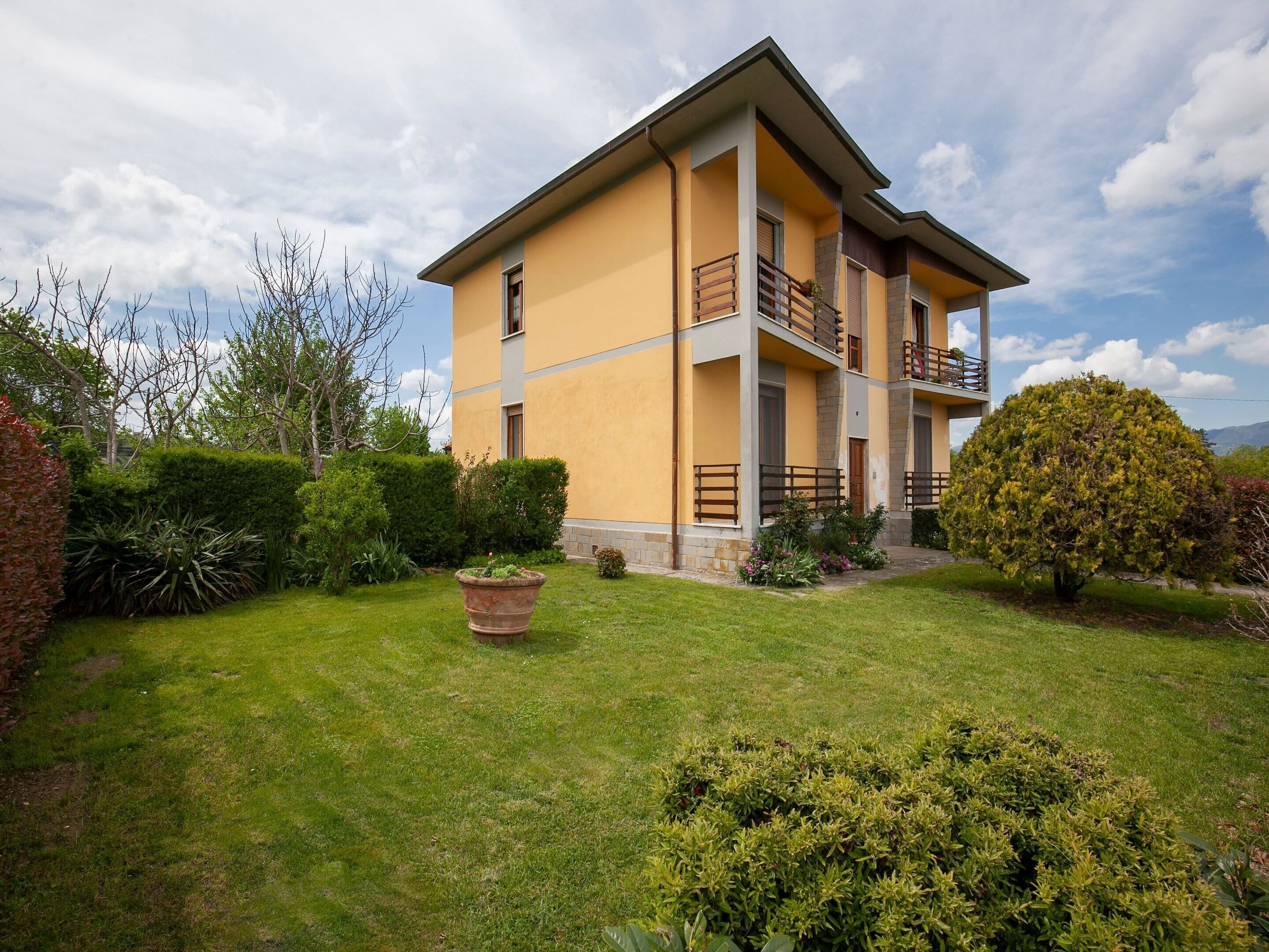 Un immeuble de deux étages avec une façade jaune, un balcon à l’étage supérieur, et une pelouse bien entretenue agrémentée d’un grand arbuste et d’une plante en pot.